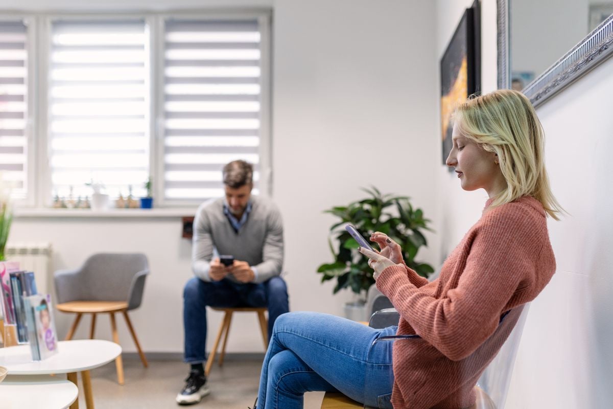  Two people sitting in a clinic waiting room, each using their phones, reflecting a modern and discreet approach to STD testing.